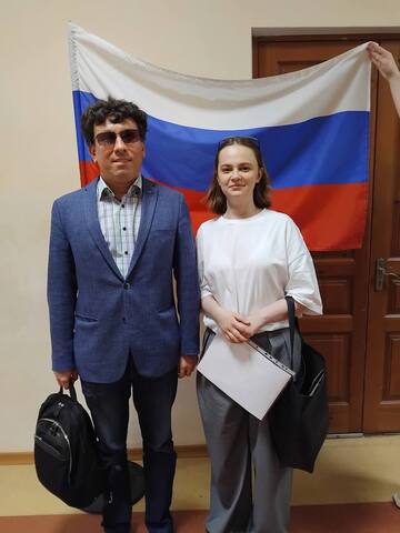 Мужчина в строгом костюме и девушка стоят с улыбками на фоне российского флага